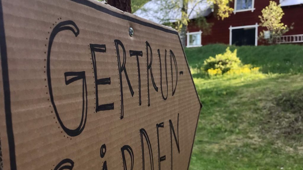 En handskriven skylt med texten Gertrudgården, som pekar på en röd byggnad.